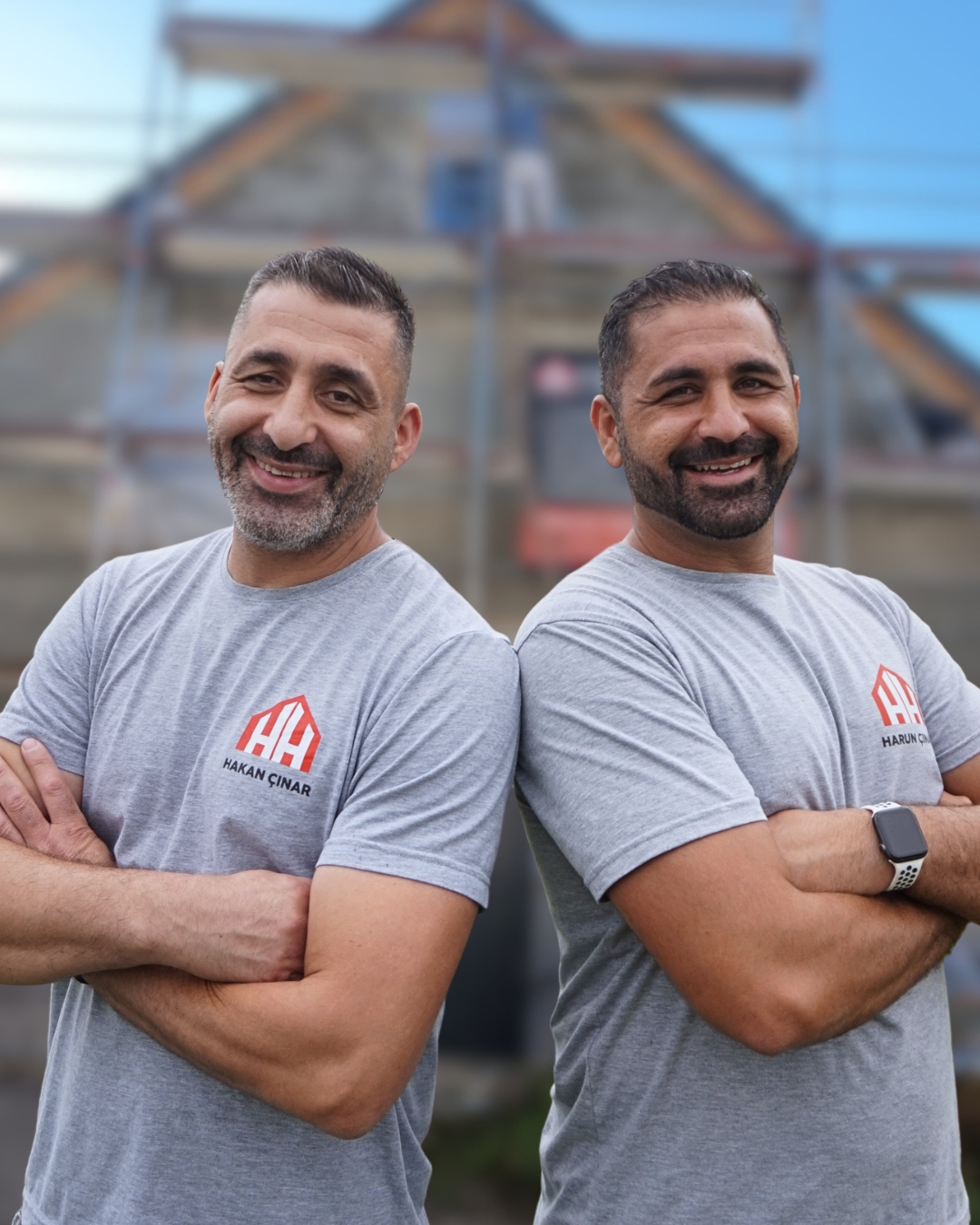 Harun und Hakan Cinar stehen mit verschränkten Armen Rücken an Rücken, beide in grauen Arbeitsshirts mit HH-Cinar-Logo