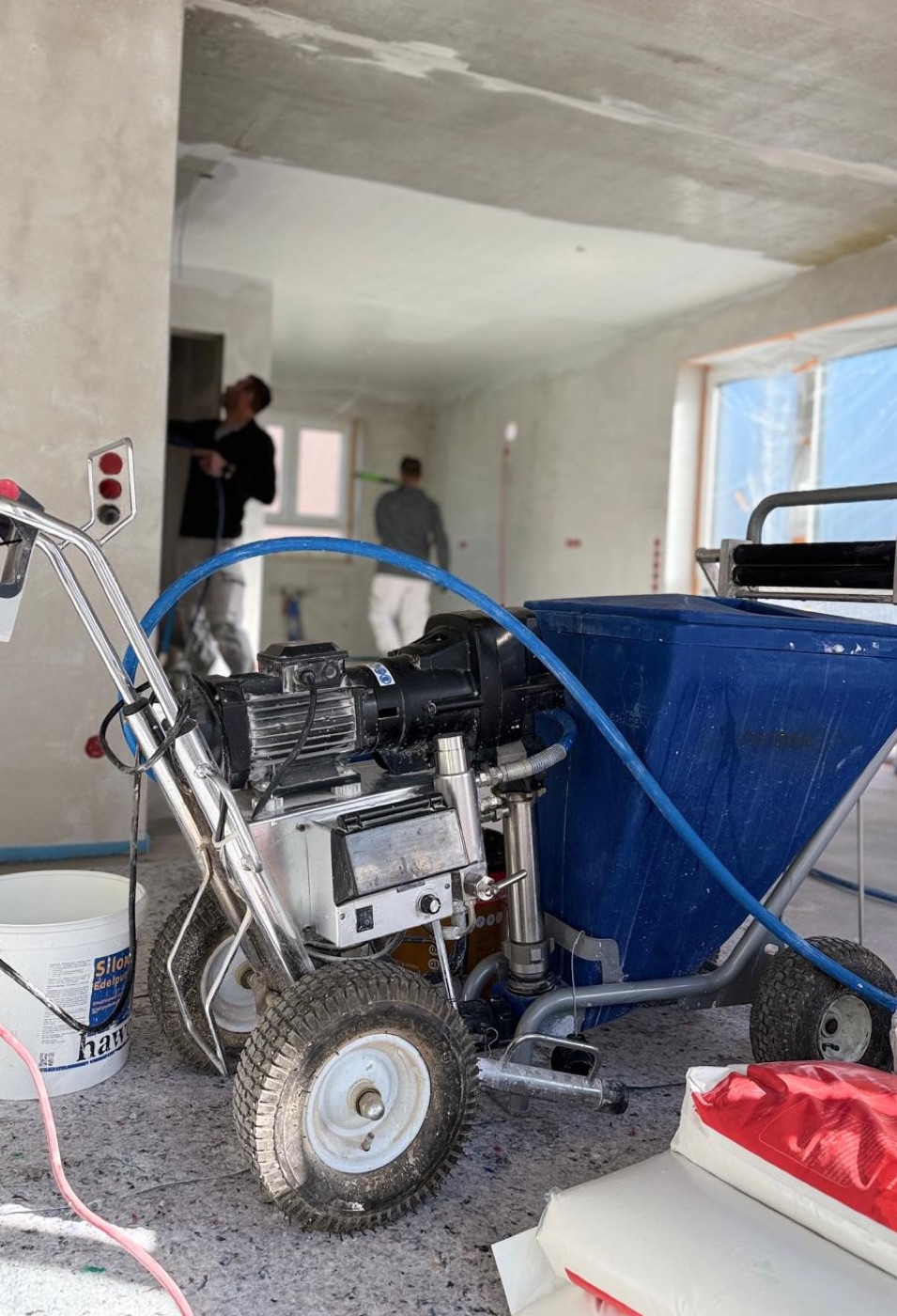 Airless-Farbspritzgerät auf der Baustelle, im Hintergrund zwei Mitarbeiter