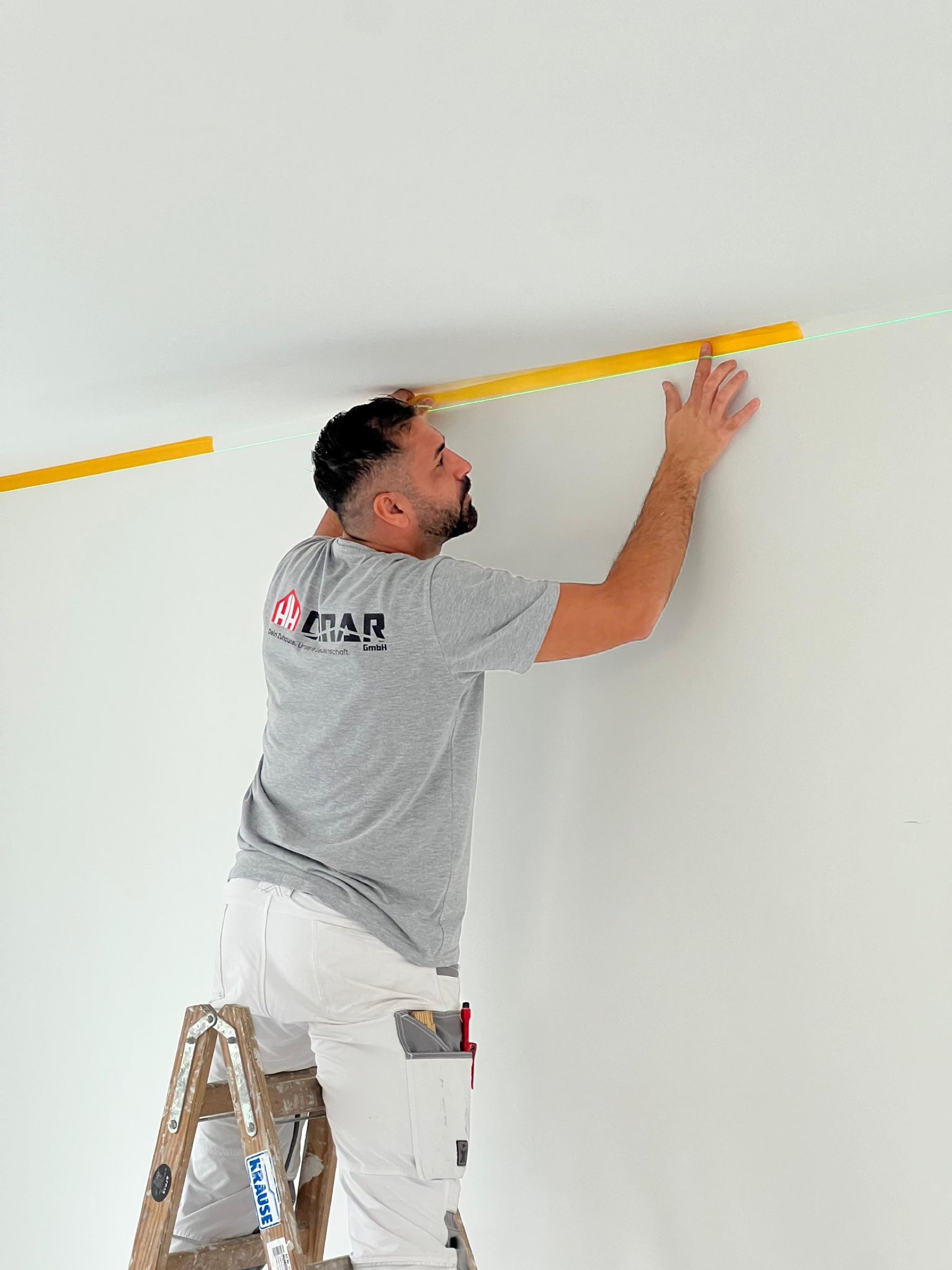 Harun Cinar klebt auf einer Leiter stehend die Kante zwischen Wand und Decke sauber ab